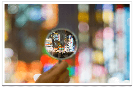 A hand holds a magnifying glass revealing a sharply focused cityscape within, against a backdrop of blurred, colorful city lights.