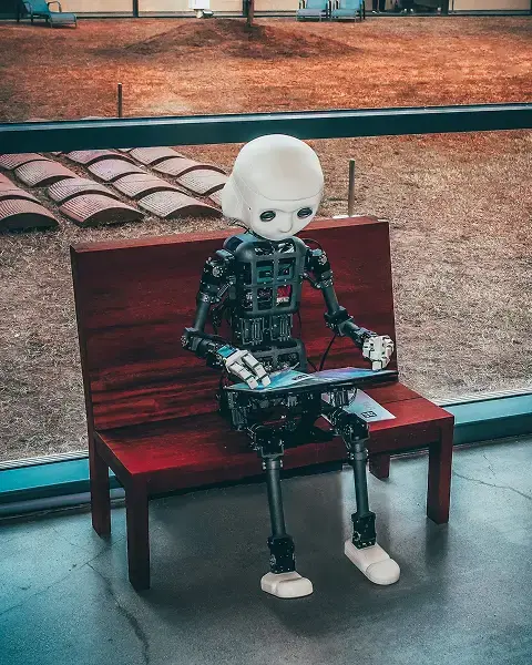 A small, humanoid robot with a white, featureless head and visible mechanical components sits on a red wooden bench, looking at a tablet-like device.