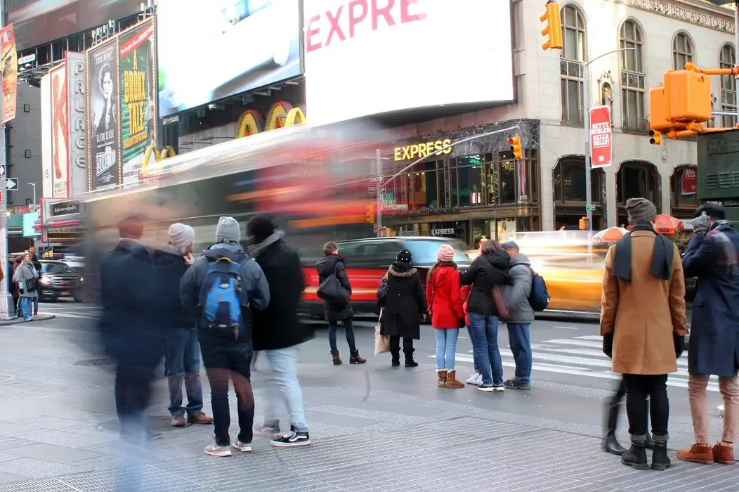 A bustling city intersection with pedestrians in winter clothing, blurred motion of vehicles, and bright storefronts and billboards in the background.